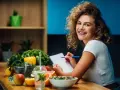 Uśmiechnięta kobieta z burzą loków planuje zdrowy posiłek, na stole świeże warzywa, owoce i woda z cytryną. Dba o dietę bez nadmiaru sodu.
