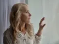 Starsza kobieta z blond włosami patrzy przez okno, zamyślona.