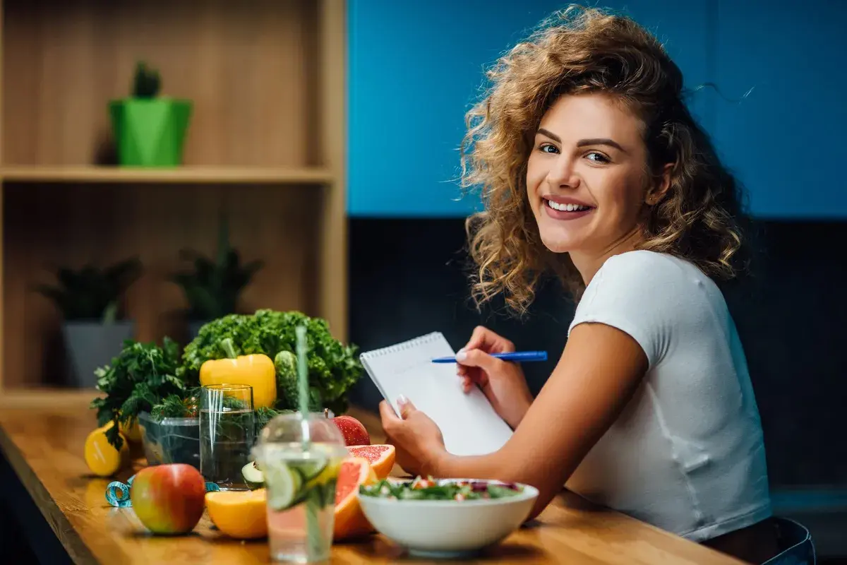 Uśmiechnięta kobieta z burzą loków planuje zdrowy posiłek, na stole świeże warzywa, owoce i woda z cytryną. Dba o dietę bez nadmiaru sodu.