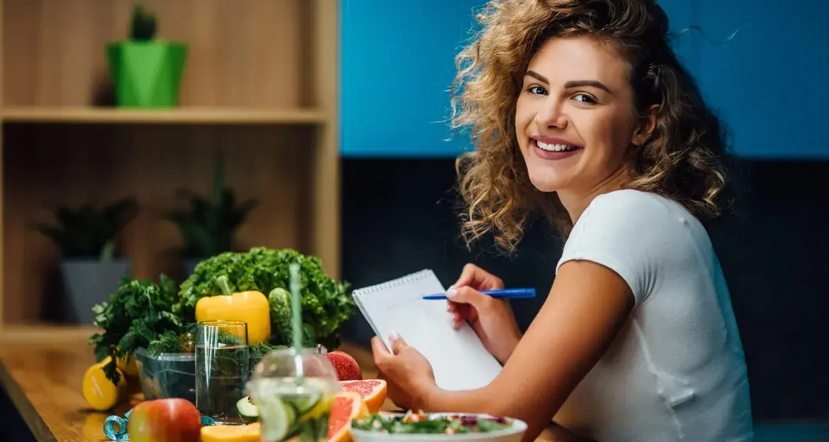 Uśmiechnięta kobieta z burzą loków planuje zdrowy posiłek, na stole świeże warzywa, owoce i woda z cytryną. Dba o dietę bez nadmiaru sodu.