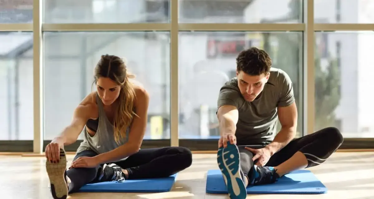Para ćwiczy stretching. Kobieta i mężczyzna siedzą na macie, wyciągając nogi i dotykając palców u stóp.