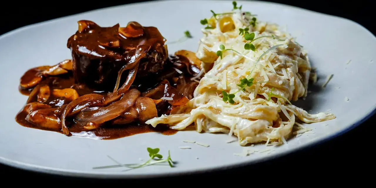 Mięso w sosie z cebulą i grzybami, przypominającymi podgrzybki, podane z kremowym makaronem posypanym parmezanem i kiełkami.