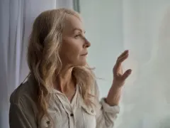 Starsza kobieta z blond włosami patrzy przez okno, zamyślona.
