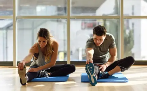Para ćwiczy stretching. Kobieta i mężczyzna siedzą na macie, wyciągając nogi i dotykając palców u stóp.