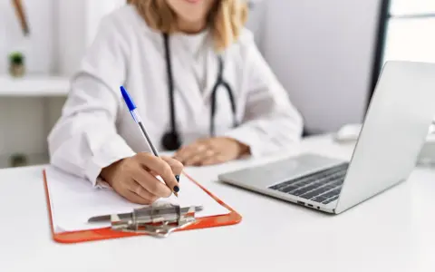Lekarka w białym kitlu pisze długopisem na kartce, obok laptop i stetoskop.