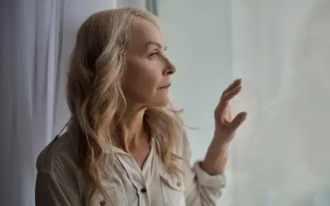 Starsza kobieta z blond włosami patrzy przez okno, zamyślona.
