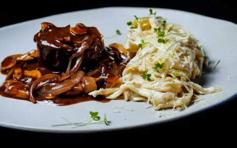 Mięso w sosie z cebulą i grzybami, przypominającymi podgrzybki, podane z kremowym makaronem posypanym parmezanem i kiełkami.