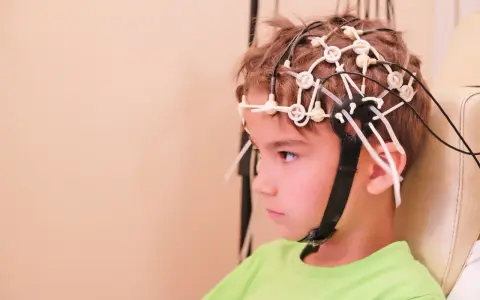 Chłopiec w zielonej koszulce ma na głowie czepki do eeg głowy, z przewodami podłączonymi do elektroencefalografu.