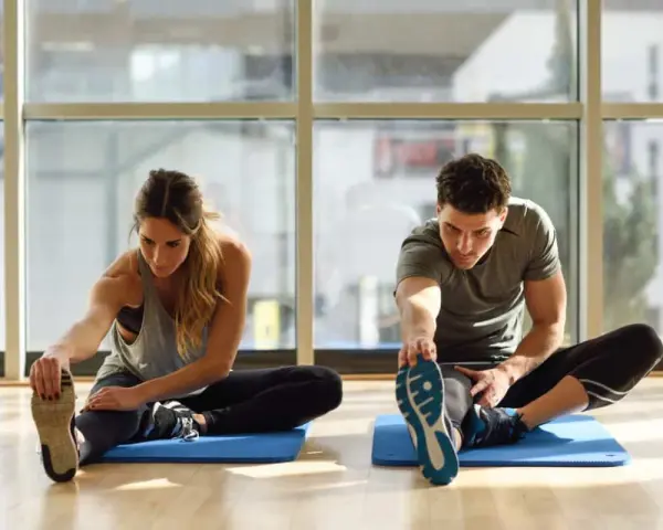 Para ćwiczy stretching. Kobieta i mężczyzna siedzą na macie, wyciągając nogi i dotykając palców u stóp.