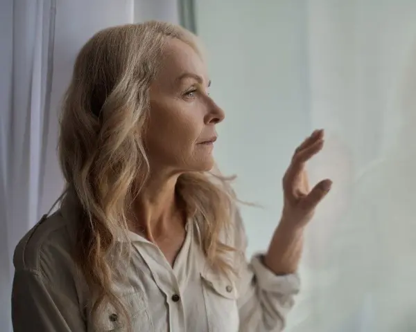 Starsza kobieta z blond włosami patrzy przez okno, zamyślona.
