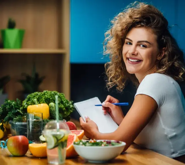 Uśmiechnięta kobieta z burzą loków planuje zdrowy posiłek, na stole świeże warzywa, owoce i woda z cytryną. Dba o dietę bez nadmiaru sodu.