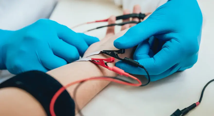Lekarz w niebieskich rękawiczkach podłącza elektrody do ręki pacjenta. To badanie EMG, które pozwala ocenić funkcje nerwów i mięśni.
