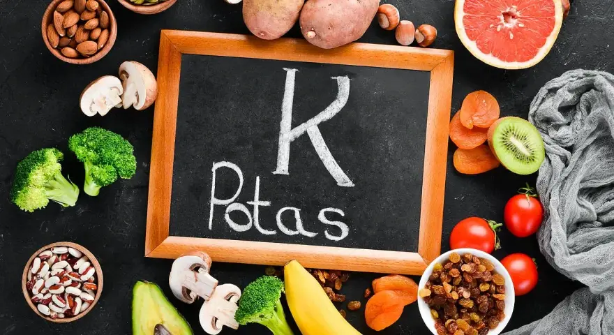 Tablica z napisem "K Potas" otoczona owocami i warzywami bogatymi w potas, jak banany, awokado, pomidory, ziemniaki, brokuły, rodzynki, morele, kiwi, migdały, pestki dyni. Pomaga zapobiegać hipokaliemii.