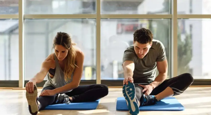 Para ćwiczy stretching. Kobieta i mężczyzna siedzą na macie, wyciągając nogi i dotykając palców u stóp.