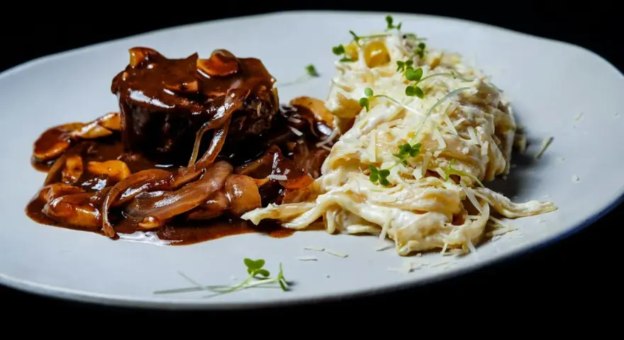 Mięso w sosie z cebulą i grzybami, przypominającymi podgrzybki, podane z kremowym makaronem posypanym parmezanem i kiełkami.
