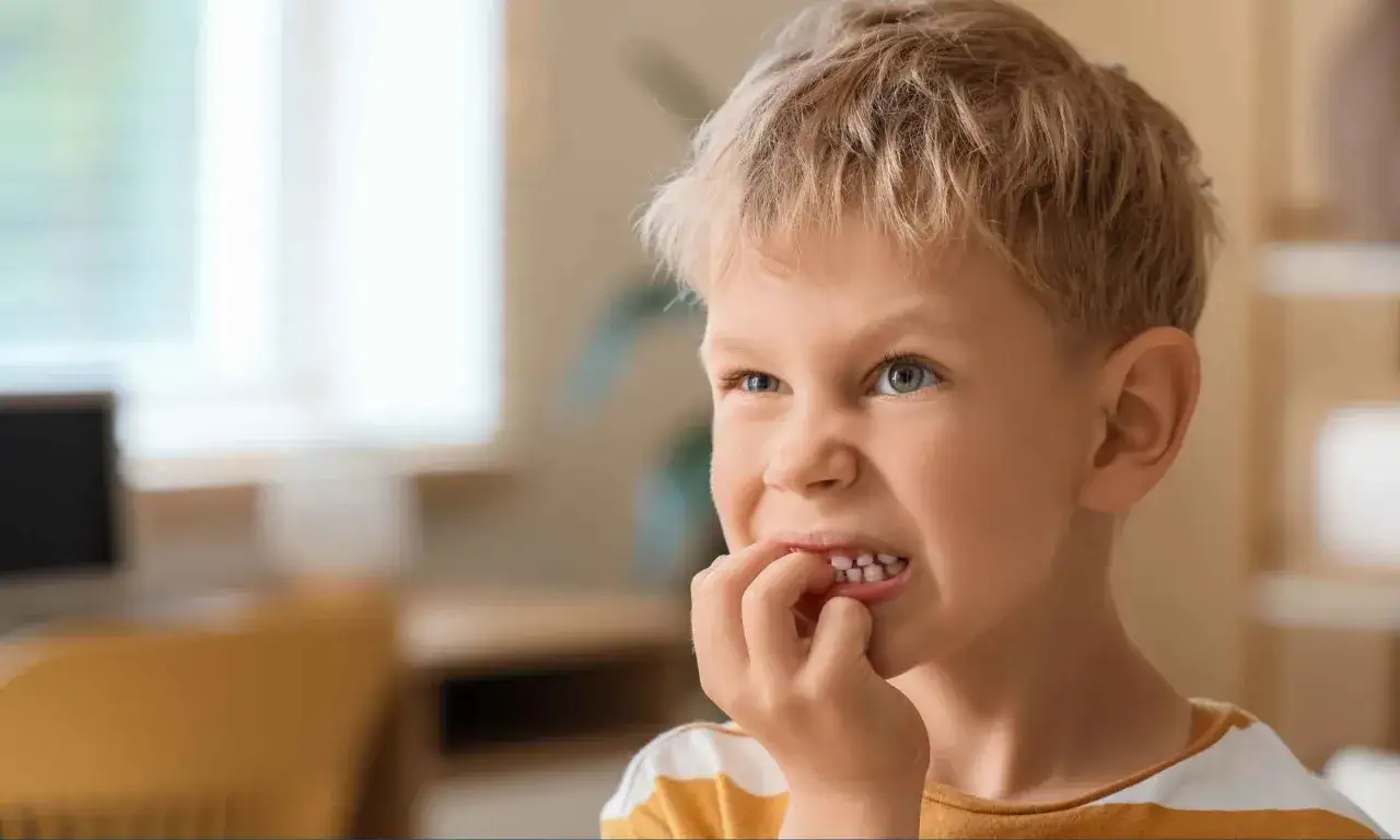 Chłopiec z blond włosami i niebieskimi oczami, który gryzie paznokcie. Może to być objaw stresu lub zespół Tourette'a.