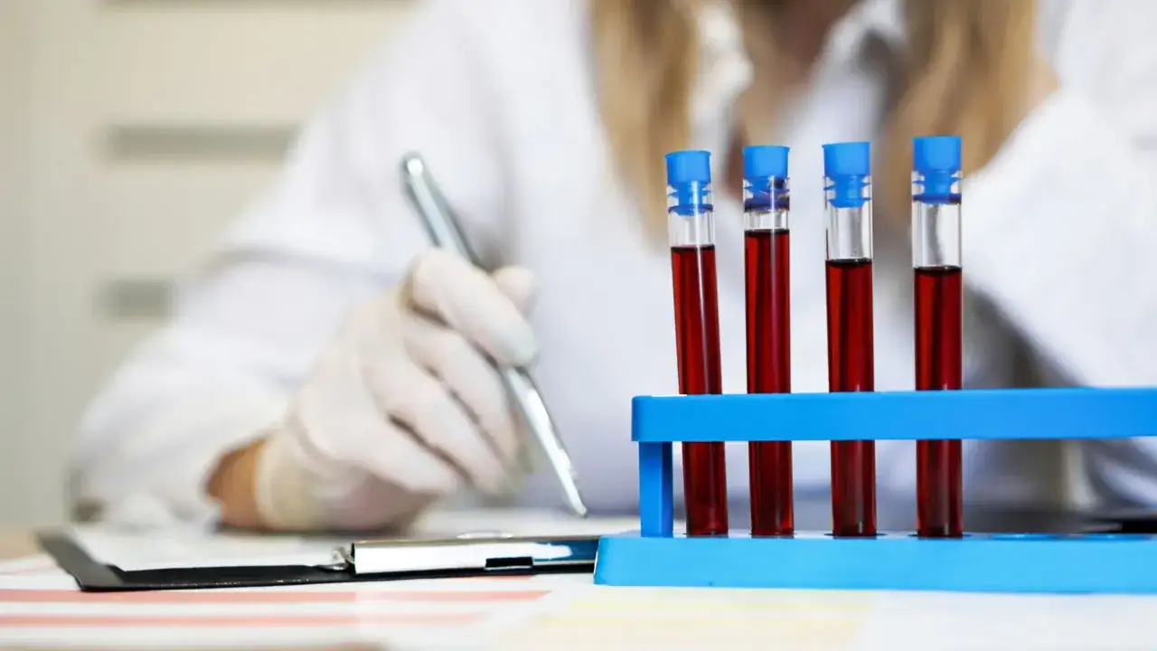W laboratorium, w probówkach z niebieskimi nakrętkami, widać krew. Ręka w białej rękawiczce pisze coś długopisem.