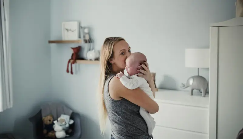 Mama tuli noworodka, by ukoić jego czkawkę. W tle pokój dziecięcy z pluszakami.